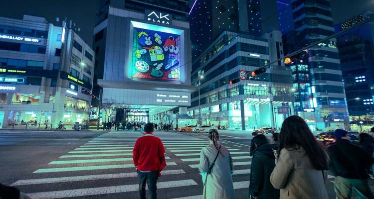 Des gens traversent un large passage pour piétons sous des lumières néon et une immense fresque numérique dans une ville coréenne la nuit.