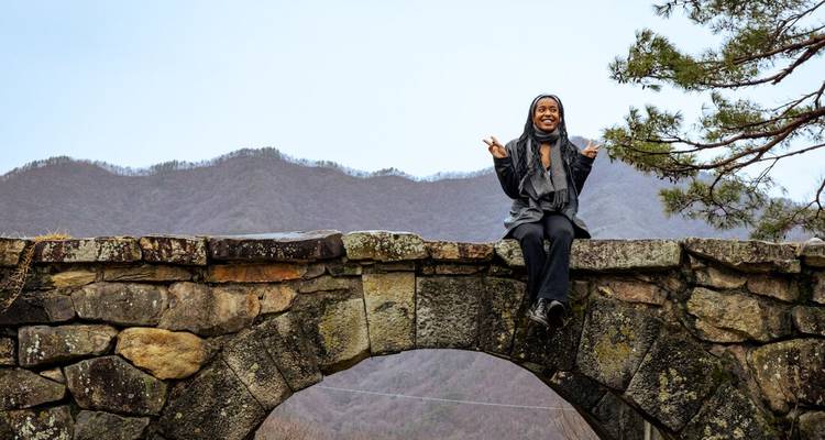 Une femme fait des signes de paix en étant assise sur un ancien pont de pierre avec une montagne en arrière-plan.
