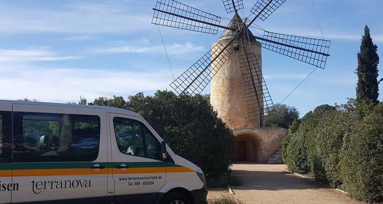 Ein traditionelle Steinwindmühle steht hinter einem Tourismusminibus, der auf einem staubigen Weg zwischen Hecken geparkt ist.