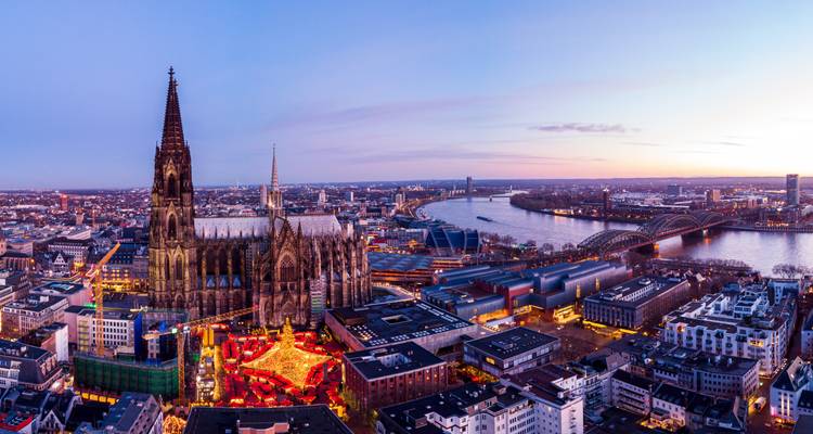 Vista aérea al atardecer de la Catedral de Colonia elevándose sobre un mercado festivo junto al río Rin.