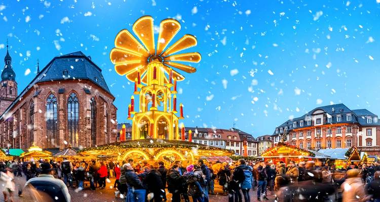 Festlicher Weihnachtsmarkt mit einem hohen beleuchteten Karussell und Schneeflocken, die über einen belebten Platz fallen.