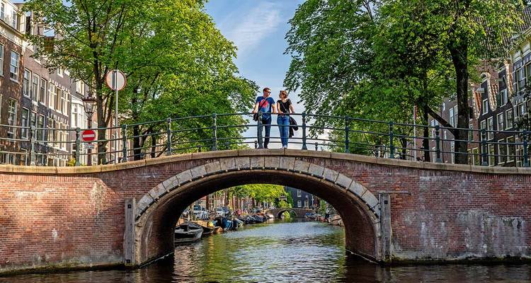 Ein Paar steht auf einer charmanten Ziegelsteinbrücke über einen Amsterdamer Kanal, der von Bäumen und historischen Häusern gesäumt ist.