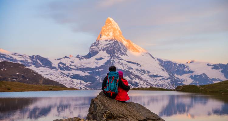 Een stel in rode jassen zit op een rots naast een alpien meer en bewondert de goudgekleurde Matterhorn in het schemerlicht.