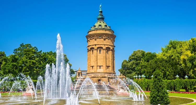 Manheims historischer Wasserturm, umrahmt von tanzenden Brunnen und üppigen Gärten unter einem strahlend blauen Himmel.