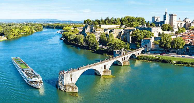 Een rivierkruisschip vaart langs de historische Pont d'Avignon over de Rhône, omgeven door groene oevers.