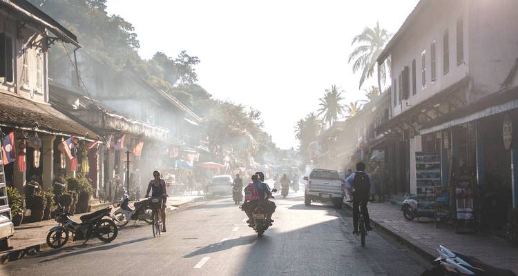 Morgenlicht und Dunst entlang einer Kolonialstraße in Luang Prabang mit Radfahrern, Motorrollern und laotischen Flaggen
