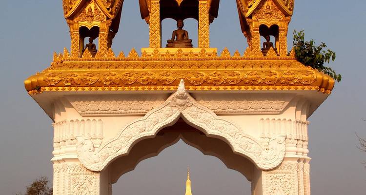 Ein verziertes goldenes Tempeltor mit aufwändigen Schnitzereien rahmt eine entfernte goldene Stupa gegen einen klaren blauen Himmel ein.