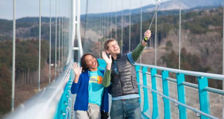 Ein lachendes Paar macht ein Selfie auf einer langen Haengebruecke ueber einem bewaldeten Tal.