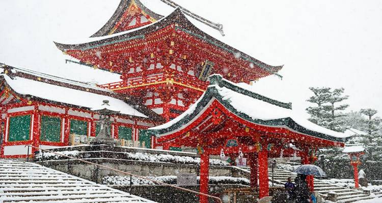 Schneefall, der die leuchtend roten Gebäude eines Kyoto-Schreins bedeckt, mit einem Besucher, der einen Regenschirm hält.
