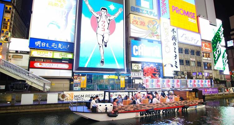 Nächtliche Ansicht des Dotonbori-Kanals mit leuchtenden Werbetafeln und einem vollbesetzten Sightseeing-Ausflugsboot mit Passagieren.