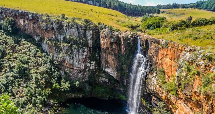 Une haute cascade se déverse sur une falaise spectaculaire de roche orange dans un bassin vert foncé, avec des collines verdoyantes et une forêt qui s'élèvent au-dessus.