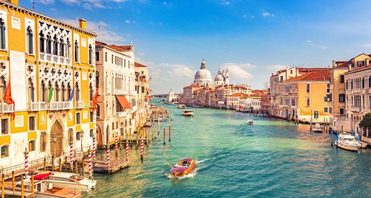 Een helder, kleurrijk uitzicht over het Groot Kanaal van Venetië met historische paleizen langs de waterweg en een klein motorbootje dat voorbijvaart; de koepels van Santa Maria della Salute in de verte.