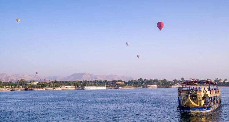 Los globos aerostáticos flotan sobre el río Nilo mientras un barco tradicional navega debajo de colinas lejanas.