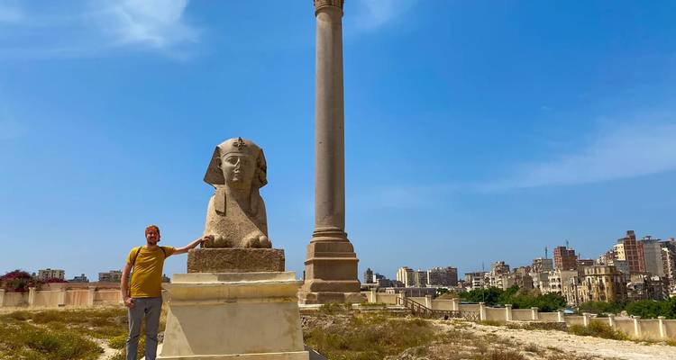 Viajero junto a una antigua estatua de la Esfinge y la Columna de Pompeyo bajo un cielo azul radiante.