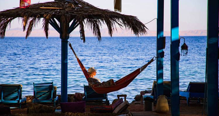 Una persona se relaja en una hamaca bajo un refugio techado con palma mirando hacia el mar tranquilo.
