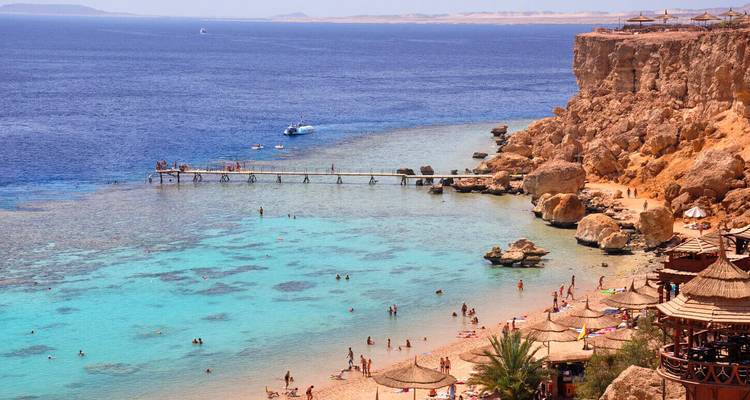 Vista desde lo alto del acantilado de una bahía turquesa con nadadores, muelle y costa desértica en un resort del Mar Rojo.