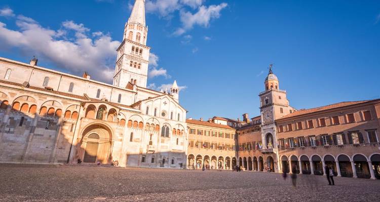 Zonnig kathedraal van Modena en omliggende plein met lange gebouwen met galerijen tegen een heldere hemel.