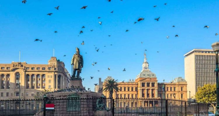 Statue dans la Place de l'Église à Pretoria avec des bâtiments gouvernementaux historiques en grès derrière et des oiseaux dans un ciel bleu dégagé