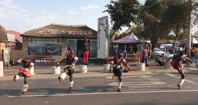 Des danseurs traditionnels effectuant des coups de pied énergiques dans une rue de township avec des étals d'artisanat et une toile de fond de huttes