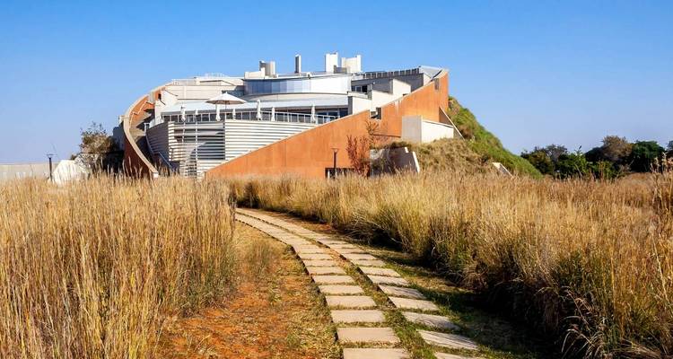 Bâtiment de musée contemporain aux murs en terre cuite courbes s'élevant au-dessus d'herbes dorées hautes le long d'un sentier en pierre