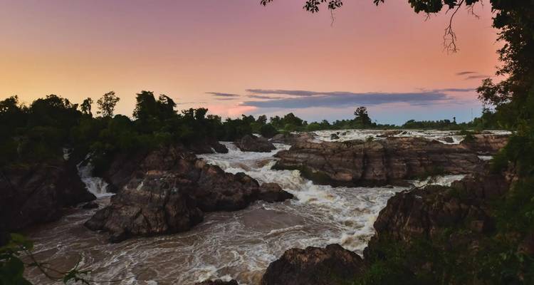 Los rápidos poderosos del río se estrellan contra rocas oscuras al atardecer bajo un cielo de puesta de sol en tonos pastel.