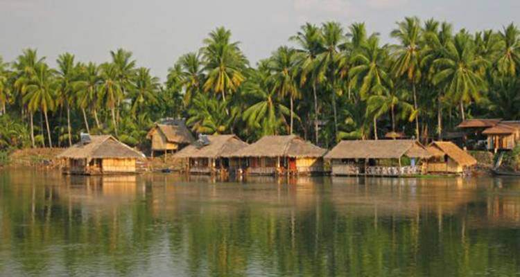 Cabañas tradicionales de paja se alinean a lo largo de una vía fluvial tranquila, con palmeras ondulantes de fondo.