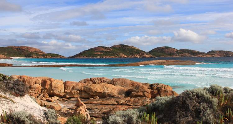 Paysage côtier rocheux avec des vagues déferlantes, des baies turquoise et des îles de granit arrondies sous un ciel partiellement nuageux.