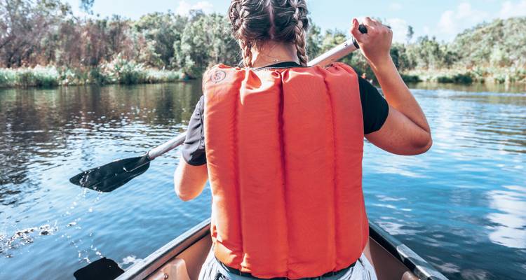 Vue arrière d'une femme pagayant dans un canoë sur une rivière calme portant un gilet de sauvetage orange.