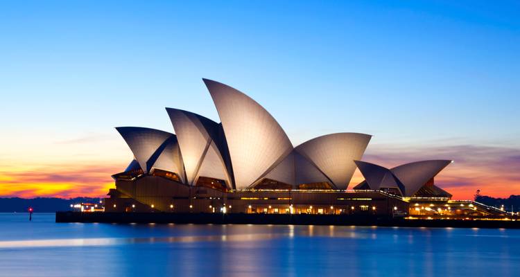 Les voiles blanches illuminées de l'Opéra de Sydney brillent contre un ciel crépusculaire coloré, se reflétant dans les eaux du port.