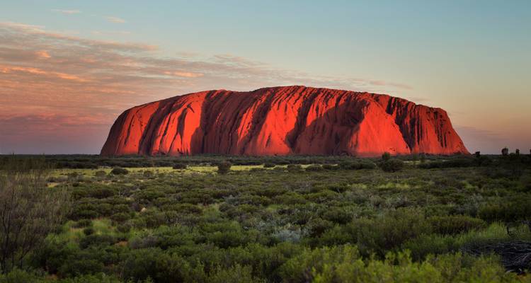 Uluru brillant d'un rouge éclatant dans la douce lumière du soir au-dessus d'une végétation désertique s'étendant au loin.