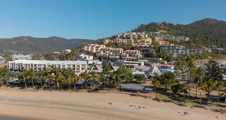 Vue aérienne de la station balnéaire côtière d'Airlie Beach avec des complexes hôteliers sur les collines, un rivage de sable et une mer bleue calme.