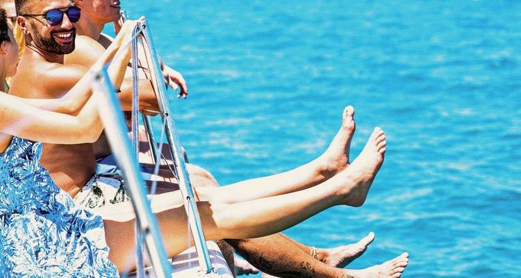 Des personnes qui se détendent sur la rambarde d'un bateau avec des jambes bronzées par le soleil qui pendent au-dessus d'une eau bleu vif.