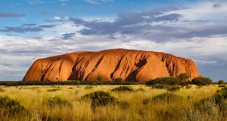 Uluru unter fleckigen Wolken, aufragend über goldenen Spinifex-Graslandschaften.