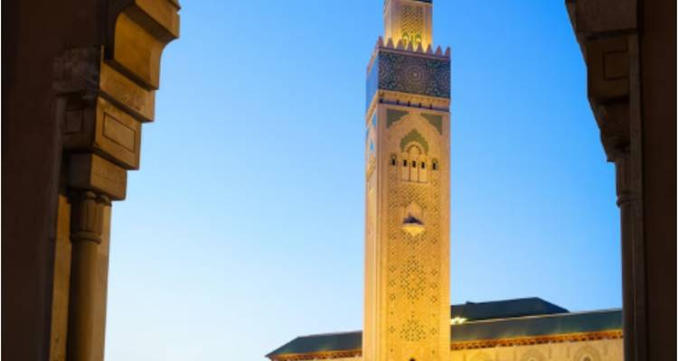 Le haut minaret de la Mosquée Hassan II encadré par une arche, illuminé contre un ciel crépusculaire.