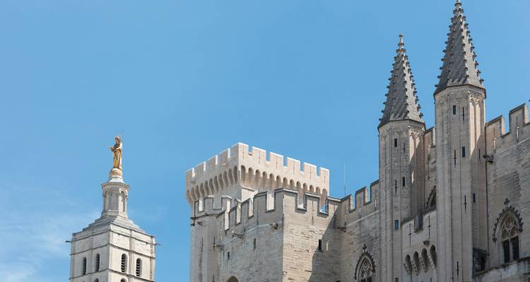 Stone spires and fortifications of the Palais des Papes rising into a clear blue sky.