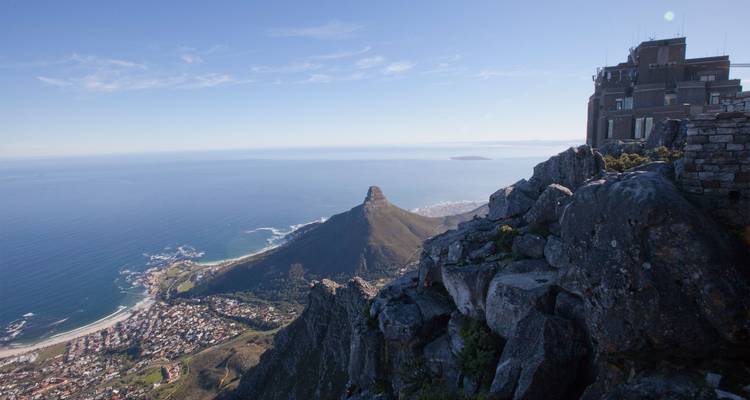 Vista panorámica desde la Montaña de la Mesa con acantilados escarpados, el pico de la Cabeza del León y la línea costera de Ciudad del Cabo encontrándose con un océano azul brillante bajo cielos despejados.