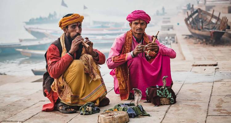 Dos encantadores de serpientes con trajes tradicionales bien coloridos están sentados con las piernas cruzadas en los ghats tocando flautas junto a sus cobras.