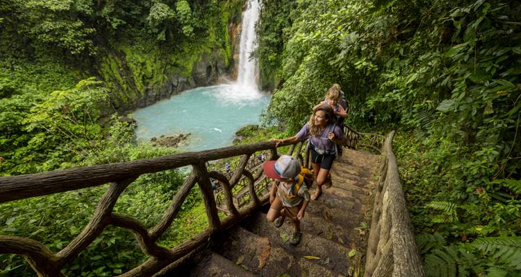 Une famille montant des escaliers en bois à travers une forêt tropicale luxuriante en direction d'un bassin de cascade turquoise au Costa Rica.