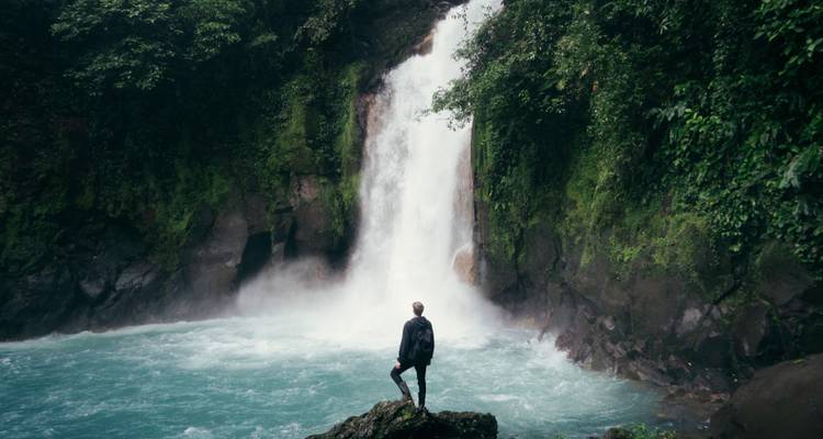 Un randonneur solitaire avec un sac à dos se tient debout sur un rocher face à une puissante cascade turquoise entourée de falaises vertes luxuriantes.