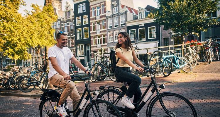 Een jong stel fietst langs een Amsterdamse straat vol geparkeerde fietsen en hoge huizen.
