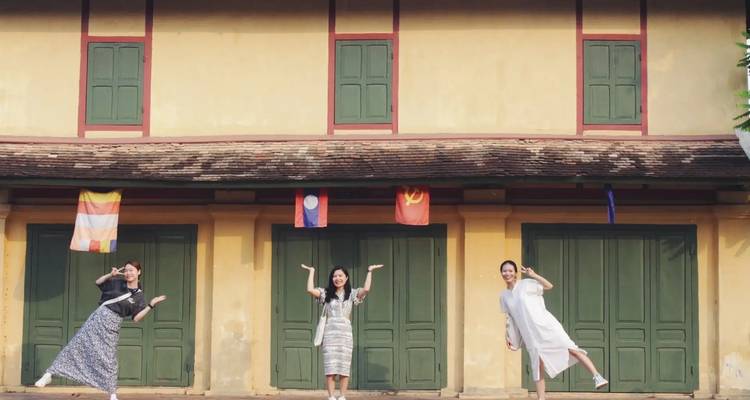 Drie jonge vrouwen in speelse poses voor een koloniaal geel gebouw met Laotiaanse en Laotiaanse vlagbanners