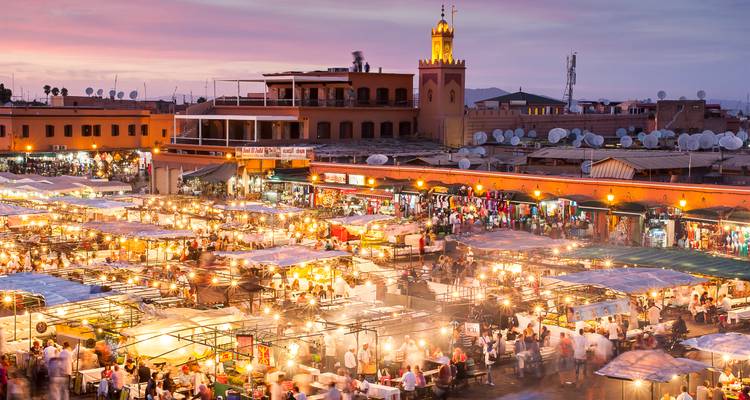 Vue en fin d'après-midi sur la place Jemaa el-Fna animée par les étals de nourriture et les lumières, avec le minaret de la Koutoubia en arrière-plan.
