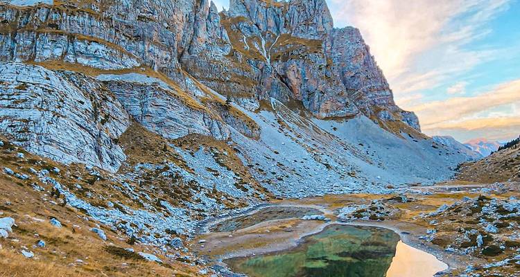 Los picos rocosos dentados se reflejan en un pequeño lago alpino bajo cielos vespertinos en tonos pastel en las Montañas Malditas.