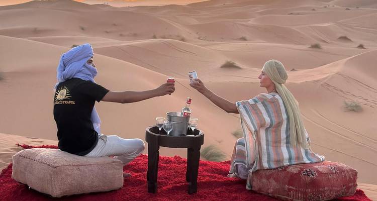 Stel zit op tapijten en stoot glazen tegen elkaar terwijl ze de zonsondergang over de rollende Sahara-duinen bekijken.