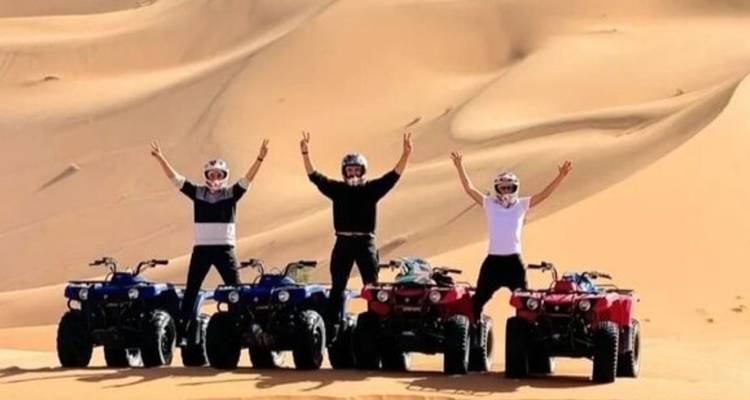 Vier ruiters steken hun armen omhoog naast quad bikes tegen torenhoge zandwanden van de Sahara.