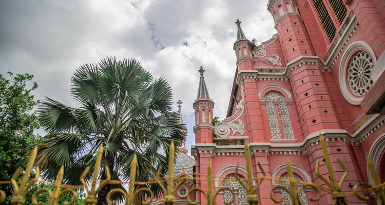 Aufwärtsblick auf die rosa Tan Dinh Kirche in Ho-Chi-Minh-Stadt, umrahmt von einer hohen Palme und bewölktem Himmel.
