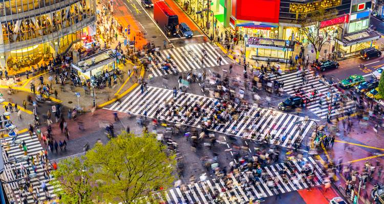 Vue en plongée du carrefour de Shibuya grouillant de piétons flous et de devantures de magasins aux enseignes lumineuses colorées.