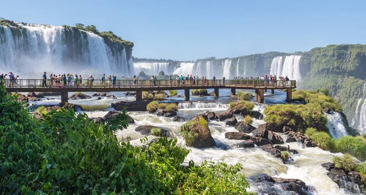 Los visitantes en un paseo observan las poderosas cascadas y la niebla de las Cataratas del Iguazú bajo cielos despejados.