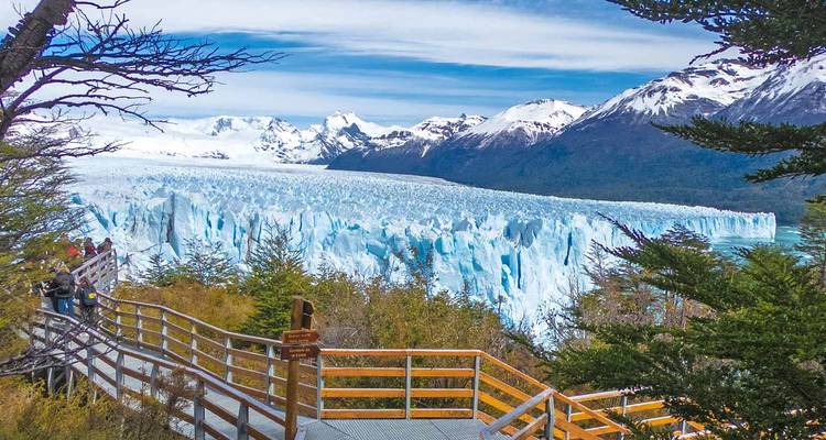 Bezoekers wandelen langs houten platforms met uitzicht op de massieve blauwe muur van de Perito Moreno-gletsjer, omlijst door sneeuwbedekte bergtoppen.