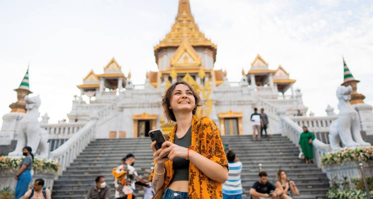 Junger Reisender, der mit einem Smartphone vor dem prunkvollen Eingang eines Bangkoker Tempels lächelt